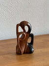 A small abstract wooden sculpture with organic curved forms in medium and dark brown tones, standing on a wooden tabletop against a textured off-white wall.