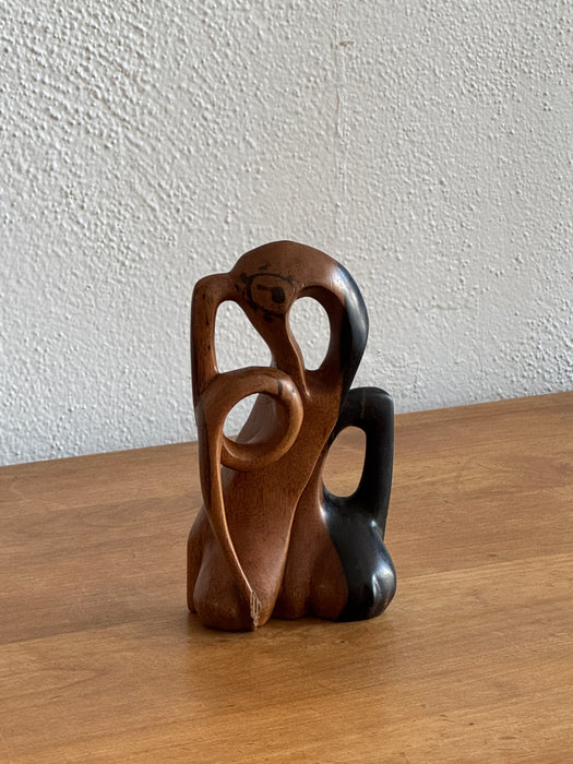 A small abstract wooden sculpture with organic curved forms in medium and dark brown tones, standing on a wooden tabletop against a textured off-white wall.