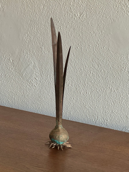 A decorative metal sculpture shaped like a bulb with tall, slender leaves, displayed on a wooden surface against a textured off-white wall.